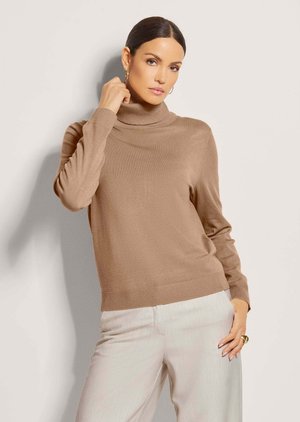 Woman with dark hair in a beige turtleneck sweater and light-colored pants, posing with one hand near her neck and the other on her hip.