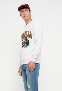 Jovem com um suéter gráfico Montana cinza-claro, boné vermelho e jeans azuis, de pé contra um fundo branco, olhando para o lado.