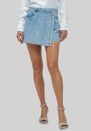 Mini-jupe en denim bleu clair avec un design enveloppant, bas effiloché et détail de bouton en métal. Associée à des sandales à talons hauts argentées à lanières.