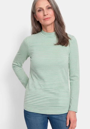 Vrouw van middelbare leeftijd met lang grijs haar die een lichtgroen-wit gestreept top met lange mouwen en blauwe spijkerbroek draagt, staand tegen een witte achtergrond.