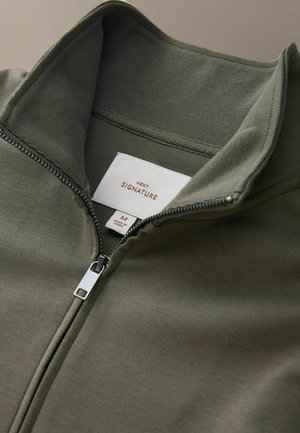 Olivovo zelená bunda na zips s vysokým golierom, vyrobená z mäkkej tkaniny, s strieborným zipsom a štítkom s nápisom "NEXT SIGNATURE".