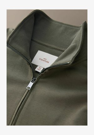 Olivgrön jacka med dragkedja och hög krage, tillverkad av mjukt tyg, med en silverfärgad dragkedja och en etikett märkt "NEXT SIGNATURE."