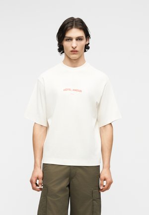 Młody mężczyzna o średniej długości ciemnych włosach, ubrany w białą, oversize koszulkę z napisem "Hôtel Amour" oraz oliwkowe spodnie cargo, stoi nieruchomo.