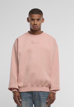 Jeune homme aux cheveux courts portant un sweat-shirt rose ample et un jean bleu, debout devant un fond gris clair uni.