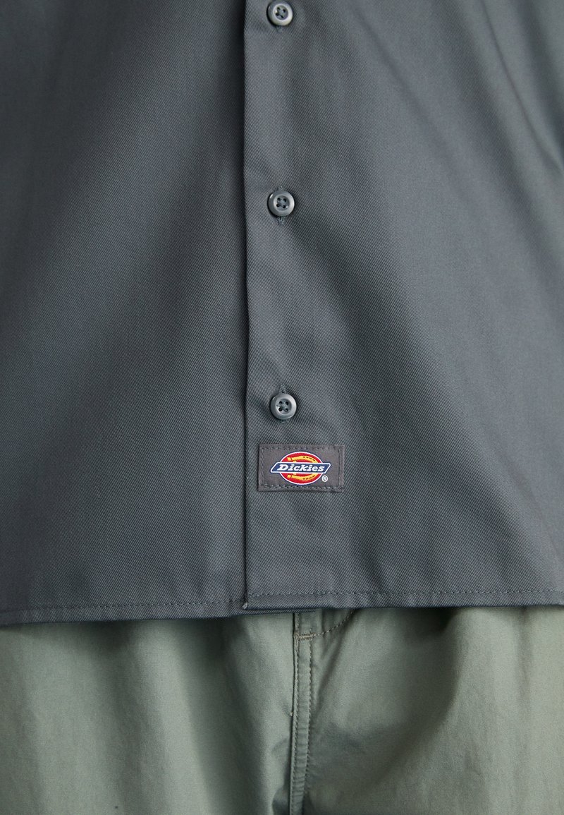 Camicia grigia con bottoni realizzata in tessuto morbido, con una piccola etichetta del logo Dickies sull'orlo inferiore. La camicia presenta bottoni scuri e un design pulito.