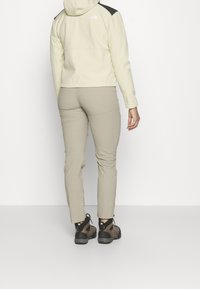 Person som bär beige vandringsbyxor, mörkbruna vandringskängor och en krämfärgad jacka med svarta lappar, står mot en vit bakgrund.