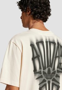 Beige bavlněné tričko s volným střihem, s velkým tmavým grafickým vzorem na zádech připomínajícím kosti a logem Adidas uprostřed.