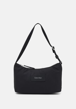 Czarna torebka na ramię Calvin Klein z regulowanym paskiem i górnym zamkiem błyskawicznym na jednolitym jasnym tle.