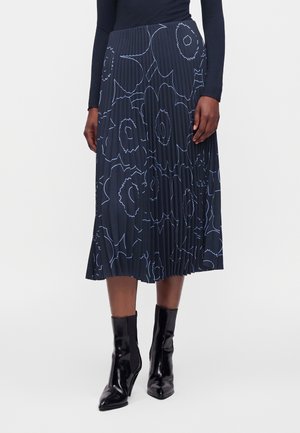 Frau trägt einen marineblauen plissierten Midirock mit großen hellblauen Blumenmustern und schwarzen Stiefeletten mit hohen Absätzen.