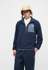 Mladý muž s béžovou zimnou čiapkou a tmavomodrou fleecovou bundou so zipsovou kapsou, stojaci s jednou rukou v kapse na jednoduchom pozadí.