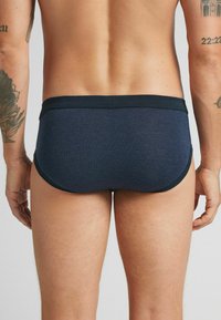 Bakifrån av en person i marinblå kalsonger med svarta detaljer, som visar tatueringar på båda överarmarna mot en enkel bakgrund.