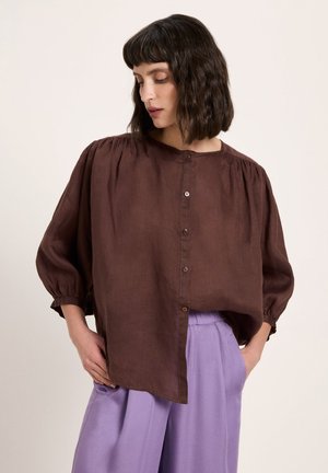 Frau mit schulterlangem, dunklem, welligem Haar trägt eine lockere braune Bluse mit Knöpfen und hellviolette, weit geschnittene Hosen und steht mit den Händen in den Taschen.