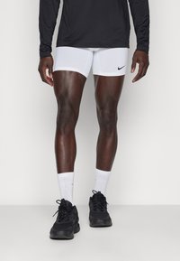 Pantalones cortos deportivos blancos con un logo negro de Nike, combinados con zapatillas negras y calcetines blancos, usados por una persona que está de pie sobre un fondo neutro.