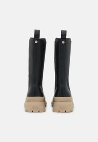 Czarne botki na kostkę z eko skóry z masywną beżową podeszwą, wyposażone w tylne pętelki i metalowe ozdoby dla dodatkowego efektu.