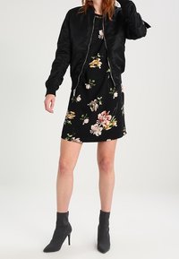 Kvinna klädd i en svart blommig klänning, svart bomberjacka och svarta ankelstövlar, stående mot en enkel ljus bakgrund.