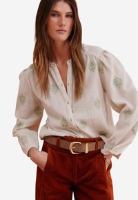 Lightweight, cream-colored blouse with green floral embroidery, button closure, and puffed sleeves, paired with rust corduroy pants and a tan belt.