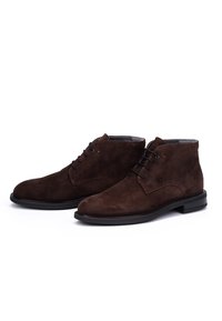 Un paio di stivali alla caviglia in suede marrone scuro con lacci neri e suole nere basse su uno sfondo bianco.