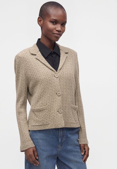 Femme portant un cardigan beige en maille torsadée avec des boutons sur une chemise noire à col et un jean bleu, debout devant un fond uni.