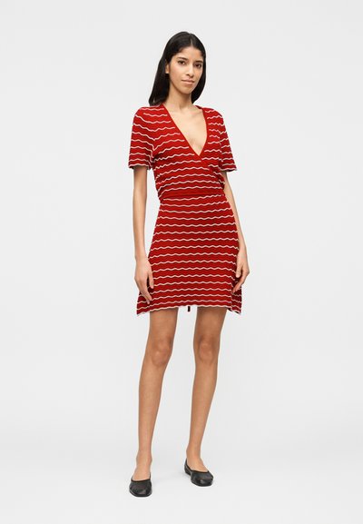 Frau steht und trägt ein rotes Wickelkleid mit weißen, wellenförmigen Streifen und schwarzen Ballerinas, posiert vor einem schlichten hellen Hintergrund.