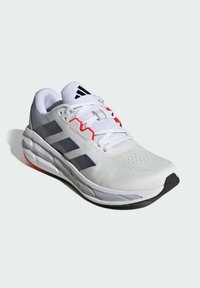 Une chaussure de sport blanche avec une tige en tissu texturé, des accents gris, des détails rouges et une semelle extérieure en caoutchouc noir. Elle présente un design à lacets.