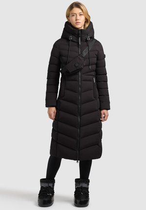 Mujer que lleva un abrigo de invierno largo negro acolchado con capucha, leggings negros y botas de invierno negras, de pie contra un fondo claro y liso.