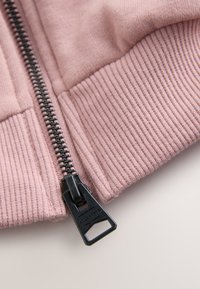 Nahaufnahme eines schwarzen Metallreißverschlusses an einem rosa gerippten Stoffrand, der den Reißverschluss-Schieber und die Strickstruktur zeigt.