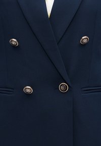 Σκούρο μπλε διπλό σακάκι με έξι διακοσμητικά χρυσά κουμπιά με πολύπλοκα σχέδια σήματος και δύο οριζόντιες μπροστινές τσέπες.