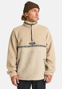 Beige fleece pullover med høj krave, halv lynlås og mønstret bånd på tværs af brystet; har sidelommer og tekstureret stof.