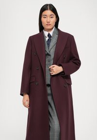 Burgunderfarbener Wollmantel mit doppelreihiger Knopfleiste, großen Revers und vorderen Taschen, getragen über einer blauen karierten Weste und einem weißen Hemd.