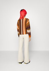 Persoon met felrood haar, gekleed in een gestreepte bruine en oranje trui, crèmekleurige gebreide broek en zwarte schoenen, staand tegen een effen achtergrond.