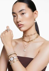 Złota kolekcja biżuterii z warstwowymi naszyjnikami, zegarkiem o srebrnej tarczy oraz wieloma błyszczącymi bransoletkami i pierścionkami z akcentami diamentowymi.