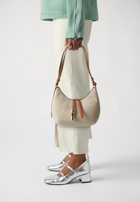 Beige textured handbag with a brown strap and gold hardware, featuring a curved shape and dual zipper closure. Silver block-heeled shoes visible.