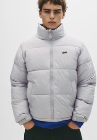 Lysegrå googlesjakke med høj krave, lynlåslukning og sort logo. Har en quiltet mønster og elastiske manchetter, båret over en blå sweater.