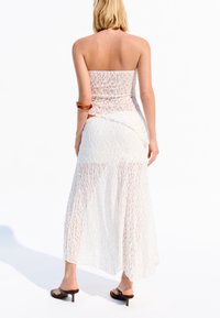 Vit off-shoulder klänning med spetsstruktur, flytande maxilängd och asymmetrisk nederkant, som är kombinerad med svarta klacksandaler och ett brunt armband.