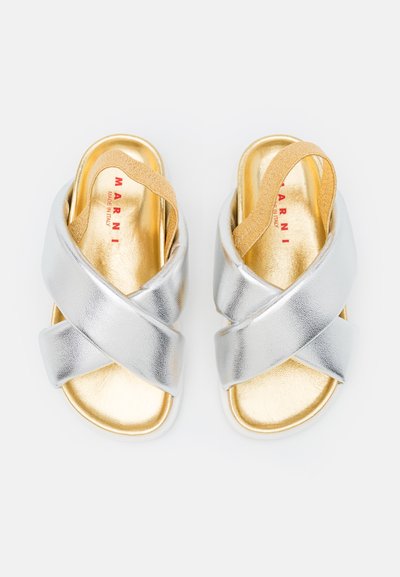 Sandali metallici argento con design a cinghia incrociata, fodera interna dorata e soletta imbottita. Presentano una cinghia elastica color sabbia per una vestibilità ottimale.