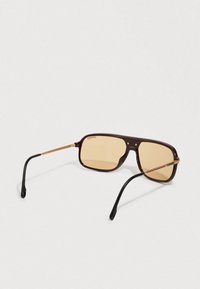 Pravougaone smeđe naočare za sunce sa jantarno obojenim staklima, tanke zlatne šake i crne zakrivljene vrhove drške koje leže na beloj pozadini.