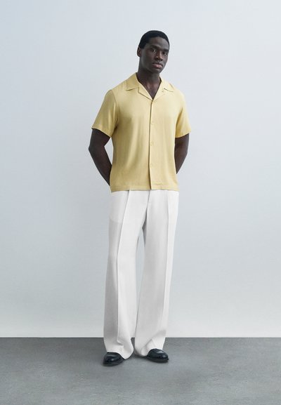 Homme debout, mains derrière le dos, portant une chemise beige à manches courtes boutonnée, un pantalon blanc à jambes larges et des chaussures noires, sur un fond uni.