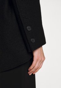 Manteau en laine noire présentant un tissu texturé et trois boutons noirs sur le poignet, avec une silhouette cintrée et une doublure intérieure lisse.