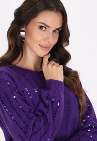 Pulover mov cu paiete, având un model în dungi, cu manșete cu riburi. Modelul pozează cu mâna pe bărbie, purtând cercei mari din argint în formă de cerc.