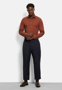 Brown button-up shirt paired with dark tailored trousers and brown dress shoes. The shirt features a classic collar and fitted design.