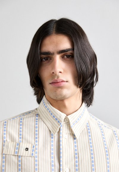 Camicia a righe beige con accenti floreali blu. Presenta un colletto, una tasca frontale e un tessuto testurizzato.