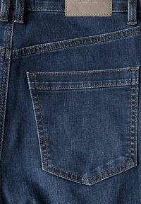 Jeans de mezclilla azul oscuro con un solo bolsillo trasero con costuras en contraste, un parche rectangular de cuero y una textura suave.