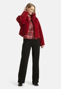 Giacca corta rossa in ecopelliccia con colletto alto, indossata sopra una maglia fantasia. Abbinata a pantaloni neri a gamba dritta e scarpe nere.