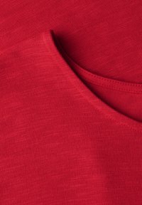 Roter Stoff mit einer glatten Textur zeigt einen runden Ausschnitt und eine kleine Tasche. Das Material ist leicht gerippt, was subtile Details hinzufügt.