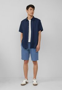 Marineblauwe kortekorte shirt met knopen, wit t-shirt eronder, blauwe shorts en witte sneakers met zwarte accenten op een model tegen een effen achtergrond.