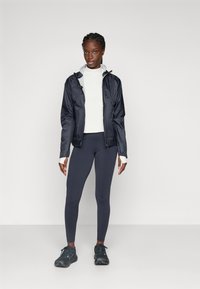Ung kvinne med flettefrisyre iført svart vindjakke, hvit langermet topp, mørke leggings og svarte joggesko som står mot en hvit bakgrunn.
