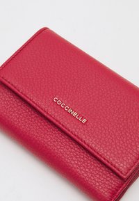 Červená kožená peněženka s texturovaným povrchem. Na přední straně se nachází zlaté logo "Coccinelle" a design s překlápěním.