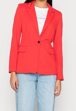 Woman wearing a bright red single-button blazer over a white t-shirt and light blue denim jeans, standing against a plain background.