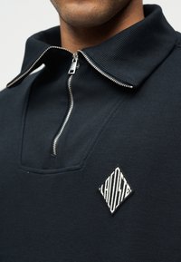 Κοντινό πλάνο ενός ατόμου που φοράει μπλε πουκάμισο με φερμουάρ και γιακά, με ένα λογότυπο σε σχήμα ρομβού, λευκό και μαύρο με υφή, στο στήθος.