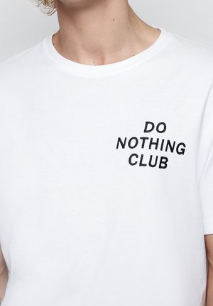 Osoba ubrana w białą koszulkę z czarnym napisem na piersi "DO NOTHING CLUB", zdjęcie kadrowane przy szyi i na wysokości środka tułowia.